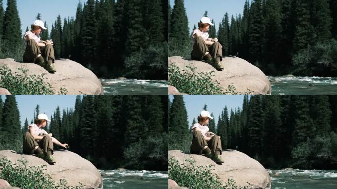 一位兴高采烈的女游客坐在山林的河上拍摄视频