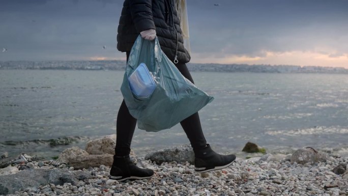 年轻的女志愿者沿着岩石海岸行走，捡起垃圾和碎片，把它们放在垃圾袋里。环保主义的概念，海滩清理，保护自