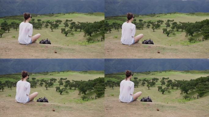 一个背包旅行的女人坐在绿色的山顶上休息，看着下面的风景