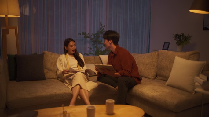 一对韩国夫妇坐在舒适的现代沙发上。年轻英俊的男子用平板电脑看新闻，而他美丽的女友正在看最新一期的时尚
