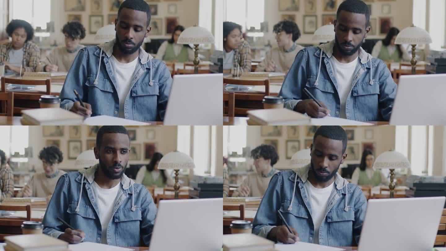 自信的非裔美国男学生在图书馆用笔记本电脑做网上研究和写笔记