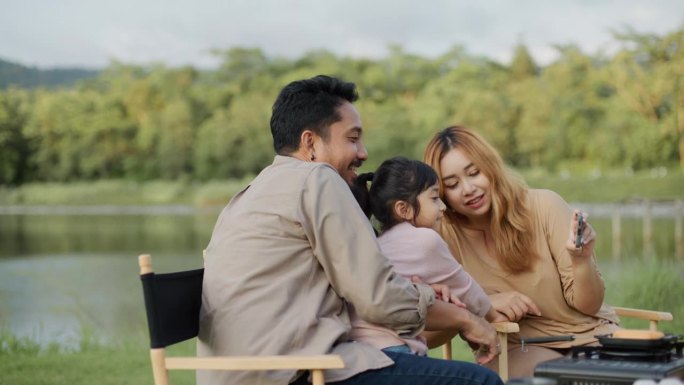 一位亚洲母亲用智能手机拍摄了女儿和丈夫一起坐在露营时的照片，并展示了照片。