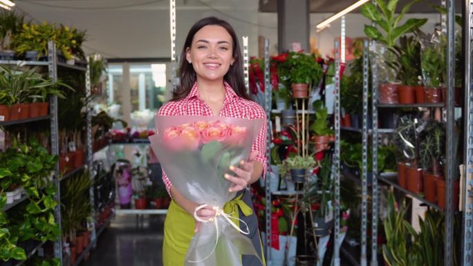 开朗迷人的年轻女花商站在花店里，手里拿着一束玫瑰花。女花商向客户卖花