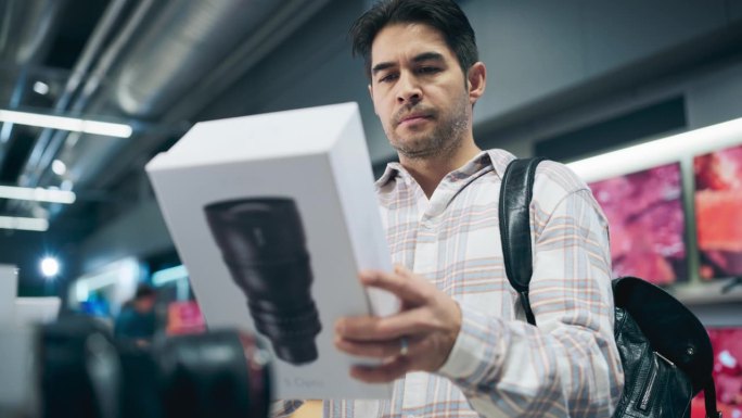 一个时髦的西班牙裔男子用现代镜头看盒子的肖像，阅读模型的规格和特征。客户在家用电器商店选购相机变焦镜