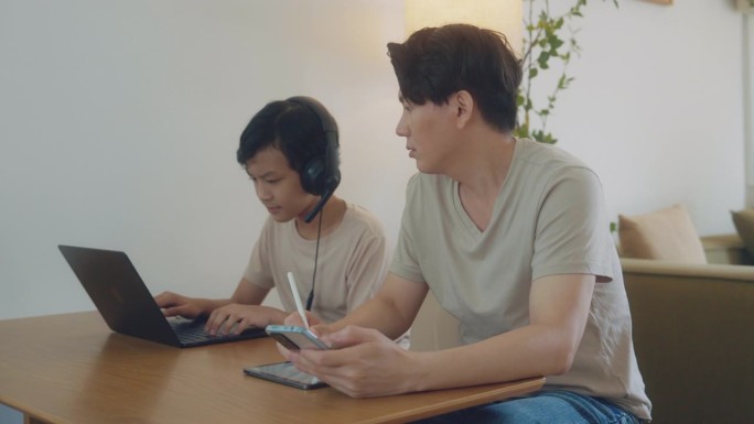 一位亚洲父亲在家工作，照顾远程在线学习外语的儿子。