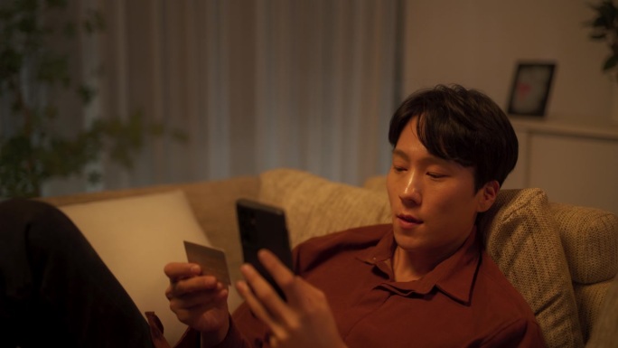 一名兴奋的韩国男子躺在舒适的客厅沙发上，用智能手机网购。亚洲男性在手机购物网站输入信用卡信息