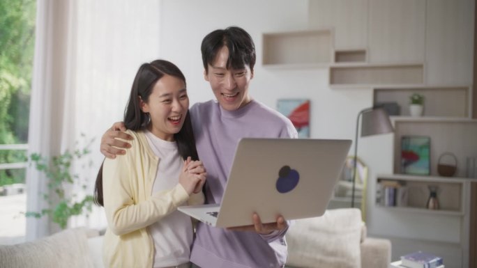 年轻可爱的韩国夫妇站在时尚的客厅里，在家里使用笔记本电脑。男人和女人等待成功的结果，快乐，欢呼和庆祝