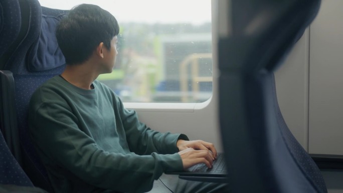 快乐的亚洲年轻企业家在火车上上下班，放松他的商务旅行，透过窗户看，在笔记本上浏览互联网。