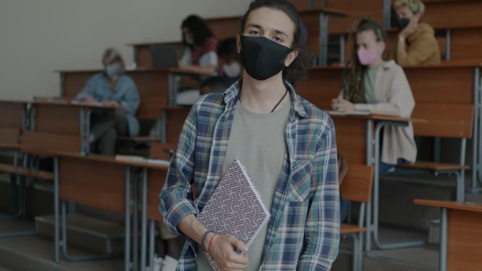 一名年轻大学生站在教室里，戴着医用口罩预防covid-19感染