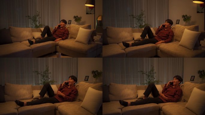 一名年轻的韩国男子正在打电话。晚上，英俊的亚洲男性舒适地坐在舒适公寓的沙发上，与朋友或家人聊天