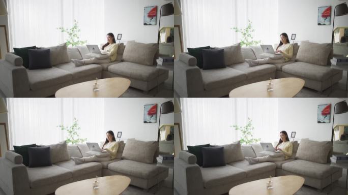 韩国女人坐在舒适的沙发上，在时尚明亮的客厅里用笔记本电脑做她的项目，与远程制作人团队合作创作项目