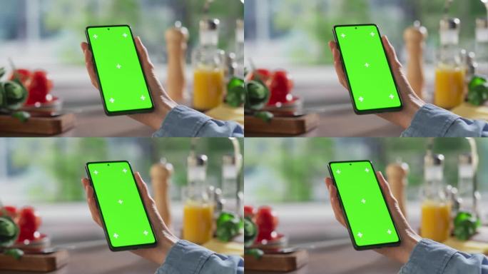 一名匿名女子手持手机，背景是厨房的桌子。智能手机绿屏色度键模板的数字营销，在线服务，应用程序，软件，