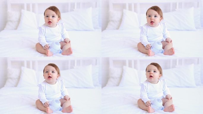 逗趣的六个月女婴穿着白色的衣服躺在床上笑着，拍着手，一个小孩子躺在家里的棉质床上，早上醒来后熟睡，概