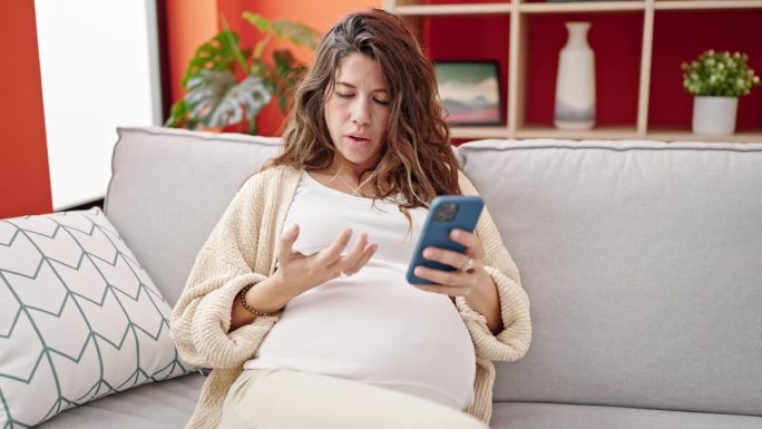 年轻的孕妇坐在家里的沙发上用智能手机和信用卡购物
