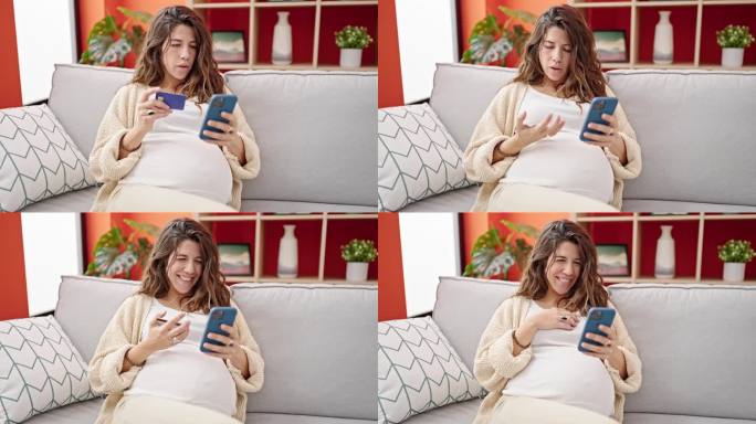 年轻的孕妇坐在家里的沙发上用智能手机和信用卡购物