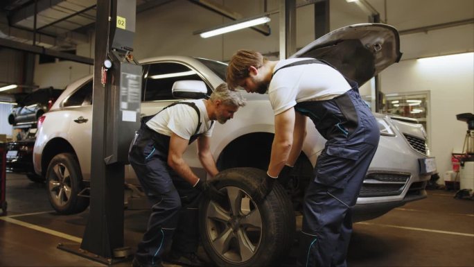 两个工人在装轮胎时安装汽车车轮。同行的机械师决定了汽车方向盘的间隙。汽车车间的工作流程。汽车车间的工
