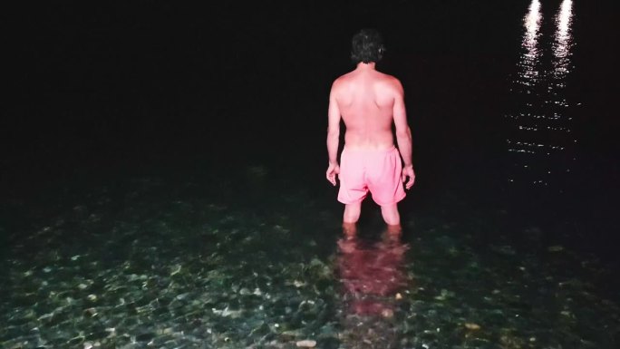 一个穿着游泳短裤的男人潜入了夜晚的大海。夜晚，在人造灯光下，清澈的海水下，卵石沙滩上五颜六色的海石