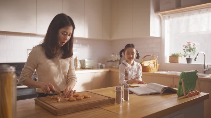 一个韩国女人和她的女儿在家里的厨房里的肖像:美丽的母亲准备做午餐，同时与她旁边的可爱的孩子交谈。幸福