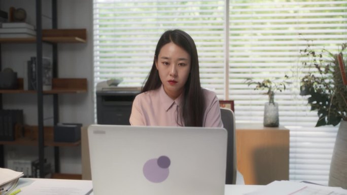 迷人的金融专家忙于分析利润，成本和收入报告在笔记本电脑上。年轻时尚的韩国专家坐在现代办公场所的办公桌