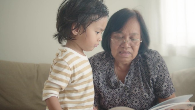 亚洲老妇人拿着一本书，她的孙子指着书页，孙子一边看书一边说话。