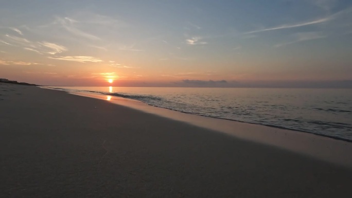 地面水平拍摄在光滑的沙滩上，因为升起的太阳的橙色光被反射在海浪中