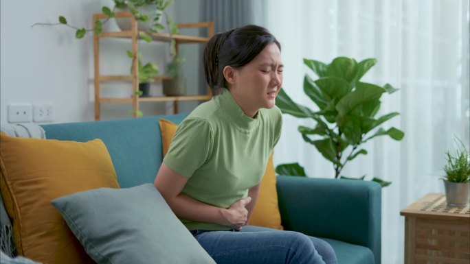 一个亚洲女人肚子疼，坐在家里客厅的沙发上。