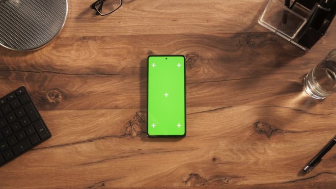 一个智能手机的顶部向下静态视图模拟绿色屏幕显示。一个手机设备躺在一个创意办公室的木桌上。在线数字营销