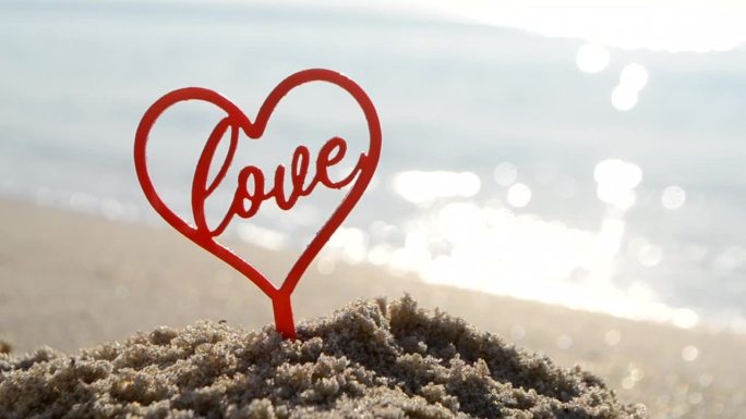 塑料棒形状的红色心脏和文字的爱在沙滩沙滩沙滩上，在阳光明媚的夏日海滨特写。人物形状的心字爱的背景海浪