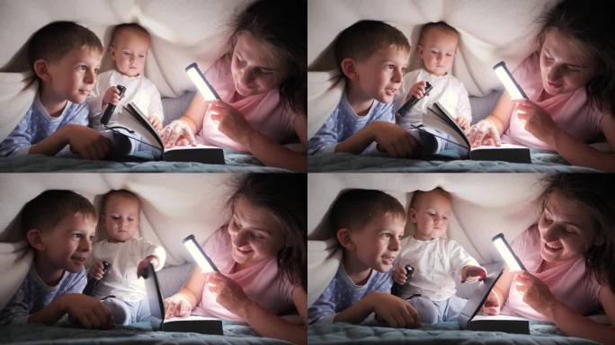 年轻慈爱的母亲给躲在被子里的两个儿子读睡前故事书。家人在一起，养育子女，快乐的童年和娱乐