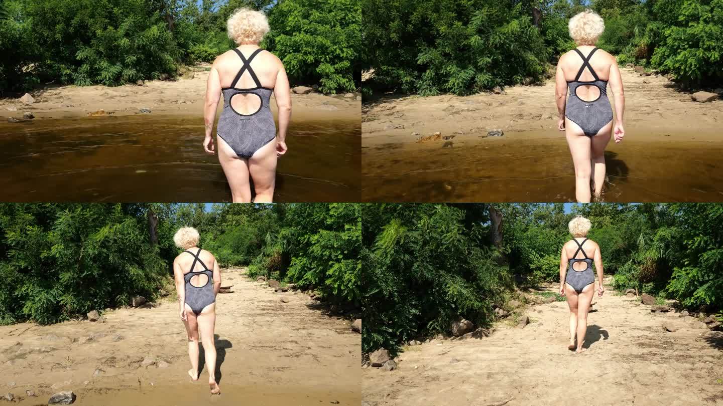 后视图一个六十岁的金发女人穿着泳衣走在水面上的海滩在丛林