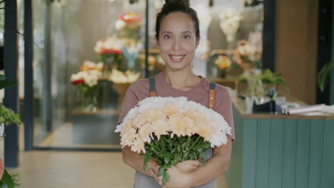 幸福的中东妇女穿着围裙站在花店，拿着新鲜的花束微笑着看着相机