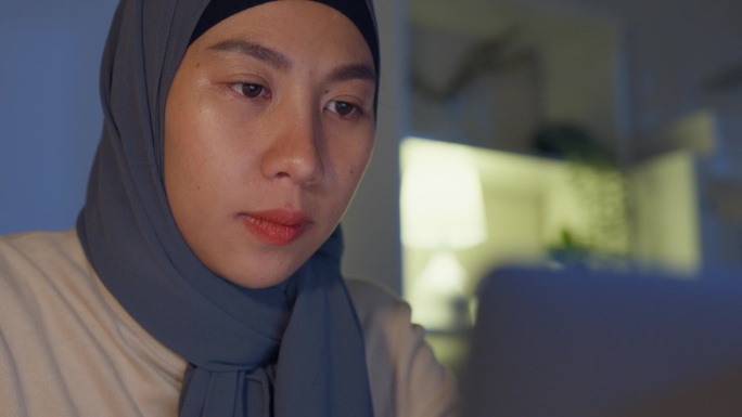 特写年轻的亚洲穆斯林自由职业者妇女戴头巾休闲布手指打字键盘笔记本电脑在家里办公室加班在晚上。小企业主