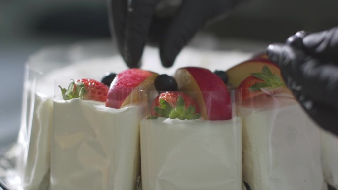 一个戴着黑色手套的厨师的手在蛋糕上把苹果片、蓝莓片和草莓片围成一个圈。装饰蛋糕。