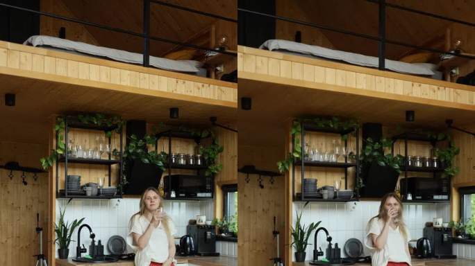 一个漂亮的女孩站在厨房里，用木房子里的杯子喝水。女人笑着离开了画框。垂直视频