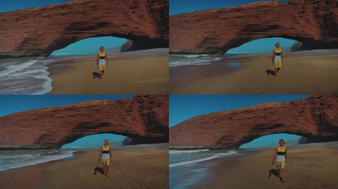 一名冲浪者腋下夹着冲浪板走在沙滩上，背景是天然的岩石拱门