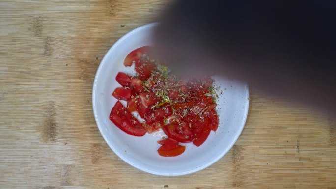 表格，俯视图，POV:一个人在一碗用橄榄油调味的西红柿中加入牛至。将牛至浇在拌有橄榄油的番茄沙拉上。