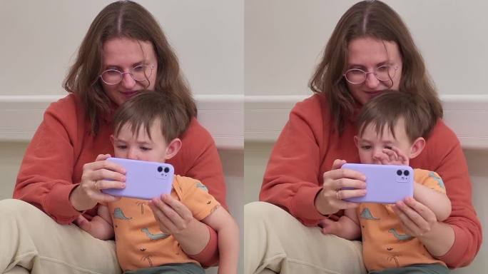 母亲和她的孩子在医院的候诊室里，在等待医生来访的时候，他们一起用手机看书。两岁男童(2岁)