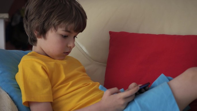 孩子坐在沙发上玩手机。男孩用手机玩电子游戏。学龄前儿童在沙发上玩电子游戏。孩子用手机玩游戏、在线教育