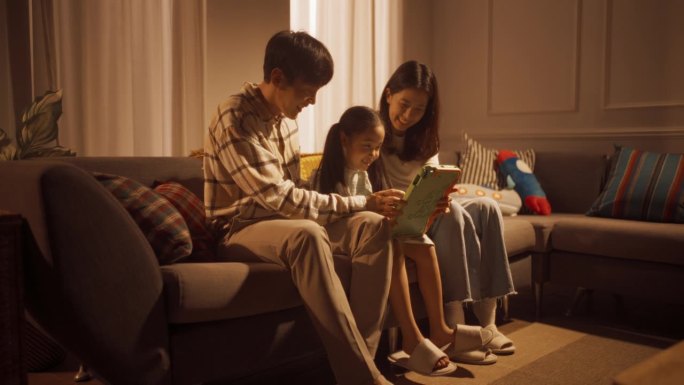 幸福的韩国家庭在家里共度时光:可爱的孩子和她的父母一起玩数码平板电脑。小女孩在慈爱的父母的照顾下享受