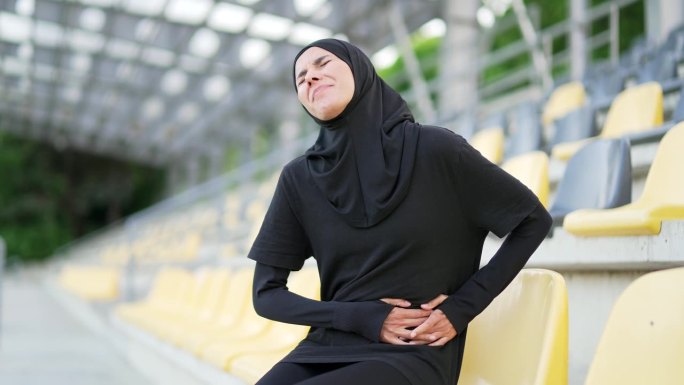 戴着头巾的年轻运动穆斯林妇女坐在城市体育场时胃痛。