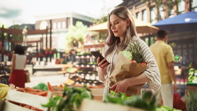 年轻漂亮的顾客购买新鲜时令水果和蔬菜，在移动中使用智能手机浏览互联网。一名妇女手持装有当地生态农产品