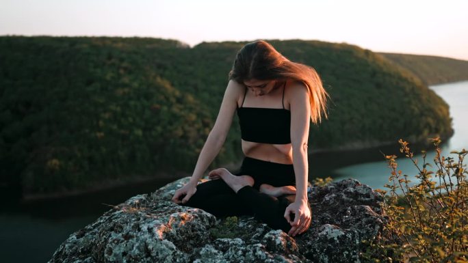 年轻女子做瑜伽腹部练习-卡帕拉巴提瑜伽呼吸技巧。瑜伽修行者在户外水面上的高悬崖上以莲花式坐着，做ud