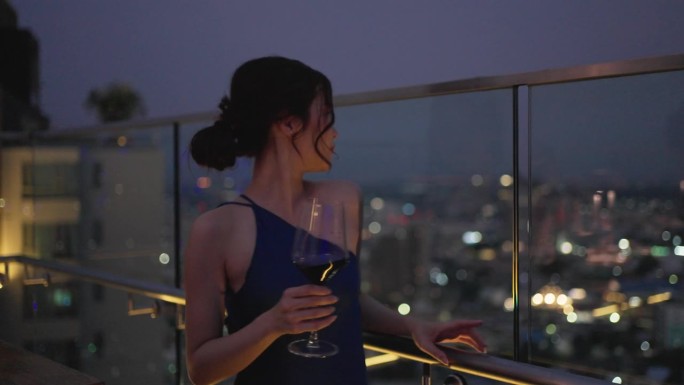 优雅的亚洲女人在城市酒店的豪华屋顶酒吧喝红酒。城市夜景。