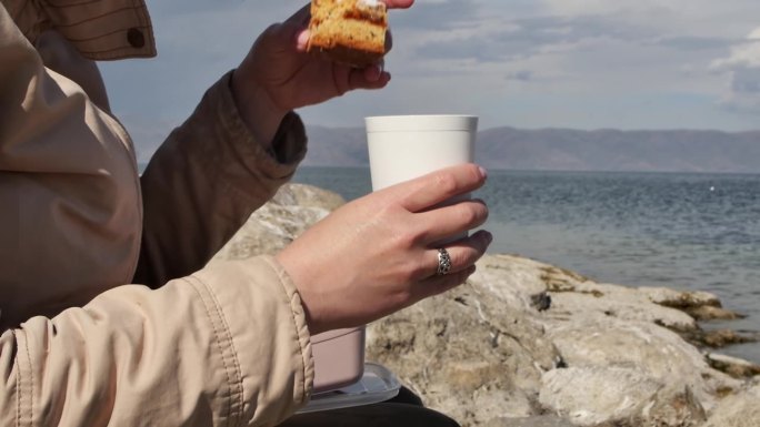 一个年轻女子坐在海边，手里拿着一杯饮料，吃着一块易碎的美味蛋糕。秋天的一天，在海边吃着甜蛋糕和热茶