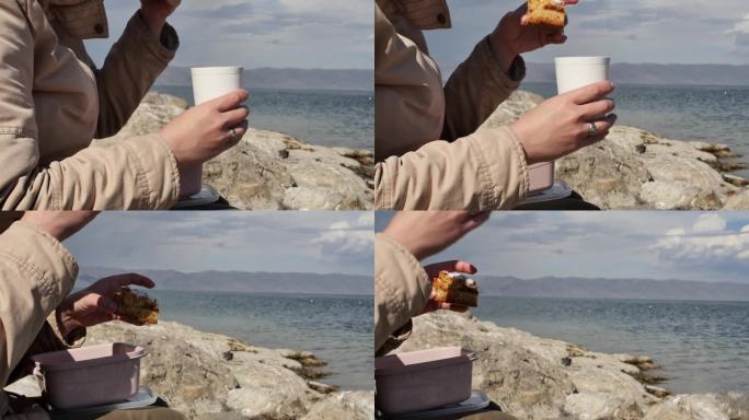 一个年轻女子坐在海边，手里拿着一杯饮料，吃着一块易碎的美味蛋糕。秋天的一天，在海边吃着甜蛋糕和热茶