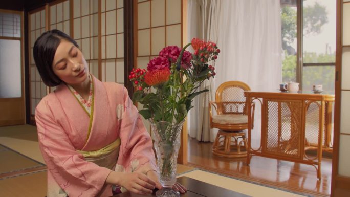 一位美丽的日本女性的肖像，穿着粉红色的和服，坐在榻榻米地板上，欣赏着一个装有彩色花束的花瓶。年轻女子