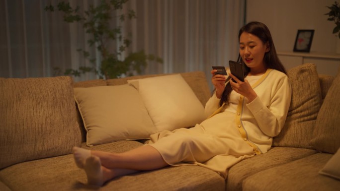 年轻的韩国女子在舒适的公寓里使用网上购物应用程序，用智能手机和信用卡安全地购物，更新流媒体服务的订阅