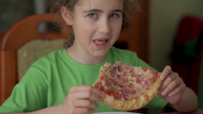饥饿的孩子咬了一口披萨。吃披萨。饥饿的小男孩坐在家里厨房的餐桌上吃着美味的意大利披萨。不健康的营养快