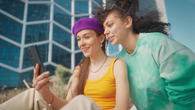 两个年轻时尚的女性朋友在城市街道上聊天，用智能手机浏览社交媒体帖子。两个时髦的女人在市中心外面讨论时