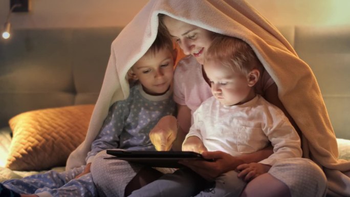 晚上盖着毯子玩平板电脑的幸福家庭。家人在一起，养育子女，快乐的童年和娱乐